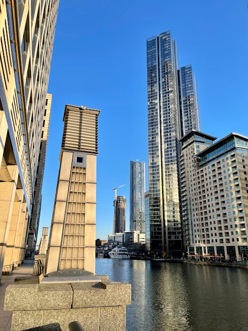 A panoramic view of the Canary Wharf skyline with modern glass skyscrapers reflecting the cloudy sky above the river. The foreground features older brick and white residential buildings alongside the